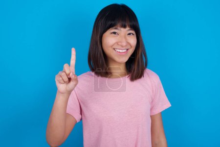 Téléchargez les photos : Jeune femme asiatique portant t-shirt sur fond bleu montrant et pointant vers le haut avec les doigts numéro un tout en souriant confiant et heureux. - en image libre de droit
