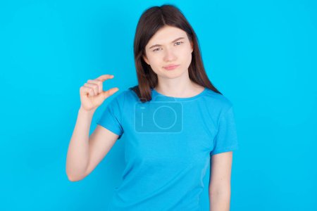 Foto de Muchacha caucásica joven descontenta usando camiseta azul aislada sobre formas de fondo azul poco signo de mano demuestra algo no muy grande. Concepto de lenguaje corporal. - Imagen libre de derechos