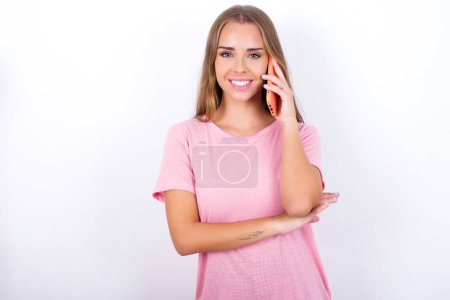 Téléchargez les photos : Portrait d'une jeune fille caucasienne souriante portant un T-shirt rose sur fond blanc parlant sur téléphone portable. Concept d'entreprise, confiance et communication. - en image libre de droit
