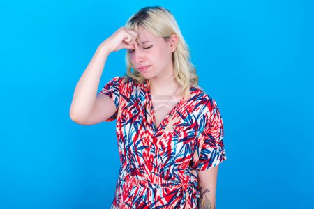 Foto de Triste chica caucásica con vestido floral aislado sobre fondo azul que sufre de dolor de cabeza sosteniendo la mano en su cara - Imagen libre de derechos
