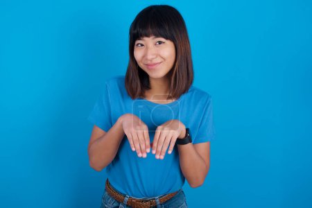 Photo for Young asian woman wearing t-shirt against blue background makes bunny paws and looks with innocent expression plays with her little kid - Royalty Free Image