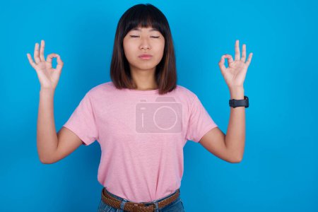 Foto de Joven mujer asiática vistiendo camiseta sobre fondo azul haciendo yoga, manteniendo los ojos cerrados, sosteniendo los dedos en gesto mudra. Meditación, religión y prácticas espirituales. - Imagen libre de derechos
