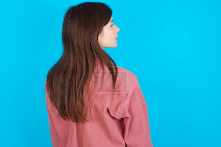 Photo for The back view of young caucasian girl wearing pink shirt isolated over blue background Studio Shoot. - Royalty Free Image