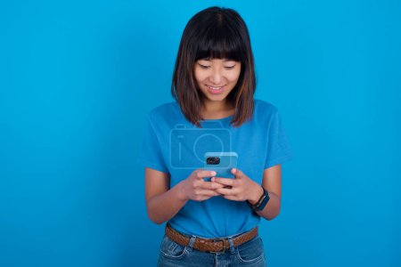 Foto de Sonriente joven mujer asiática usando camiseta sobre fondo azul usando el teléfono celular, mensajería, estar feliz de enviar mensajes con amigos, mirando el teléfono inteligente. Tecnologías modernas y comunicación. - Imagen libre de derechos