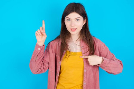 Photo for Young caucasian girl wearing pink shirt isolated over blue background says: wow how exciting it is, has amazed expression, indicates something. One hand on her chest and pointing with other hand. - Royalty Free Image