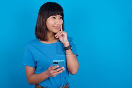 Téléchargez les photos : Pensant rêver jeune femme asiatique portant t-shirt bleu sur fond bleu à l'aide d'un téléphone mobile et tenant la main sur le visage. Prendre des décisions et concept de médias sociaux. - en image libre de droit