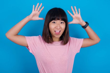 Téléchargez les photos : Jeune femme asiatique portant rose t-shirt sur fond bleu regarde avec excitation à la caméra, garde les mains levées sur la tête, remarque quelque chose d'inattendu, réagit à des nouvelles soudaines. - en image libre de droit