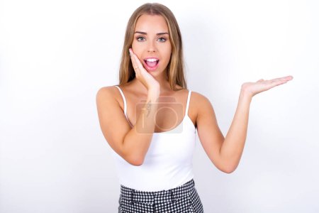 Photo for Positive glad Young Caucasian girl wearing white tank top on white background says: wow how exciting it is, indicates something.  One hand on his head and pointing with other hand. - Royalty Free Image