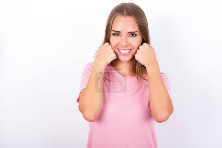 Heureuse jeune fille caucasienne portant un t-shirt rose sur fond blanc garde les poings sur les joues sourit largement et a une expression positive étant de bonne humeur