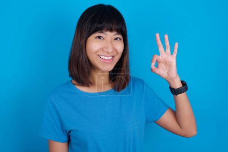 Téléchargez les photos : Jeune asiatique femme porter bleu t-shirt sur fond bleu tenir main bras okey symbole toothy approuver conseiller nouveauté nouvelles - en image libre de droit