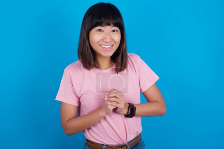 Photo for Happy young asian woman wearing pink t-shirt against blue background stands against orange studio wall keeps hands on heart, swears be loyal, expresses gratitude. Honesty concept. - Royalty Free Image