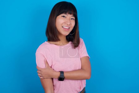 Foto de Gente, estilo de vida, juventud y concepto de felicidad. tímido bastante joven asiático mujer usando rosa camiseta contra azul fondo sensación feliz abrazándose. - Imagen libre de derechos