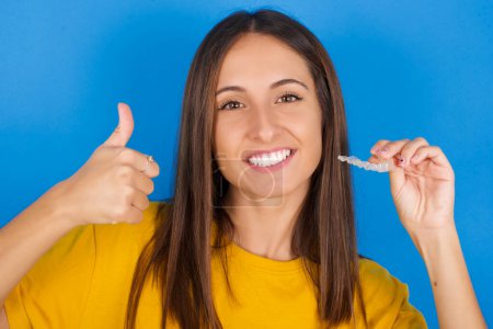 Photo for Young woman holding an invisible braces aligner and rising thumb up, recommending this new treatment. Dental healthcare concept. - Royalty Free Image