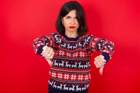 Foto de Morena mujer caucásica vistiendo suéter de Navidad sobre fondo rojo siendo molesto mostrando el pulgar hacia abajo con dos manos. Concepto de aversión. - Imagen libre de derechos
