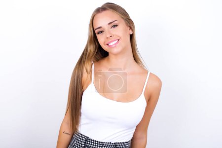 Photo for Young Caucasian girl wearing white tank top on white background with broad smile, shows white teeth, feeling confident rejoices having day off. - Royalty Free Image