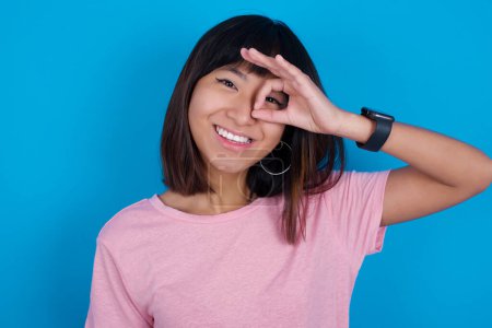 Foto de Joven asiático mujer usando rosa camiseta contra azul fondo con feliz cara sonriendo haciendo ok signo con mano en ojo mirando a través de dedo. - Imagen libre de derechos