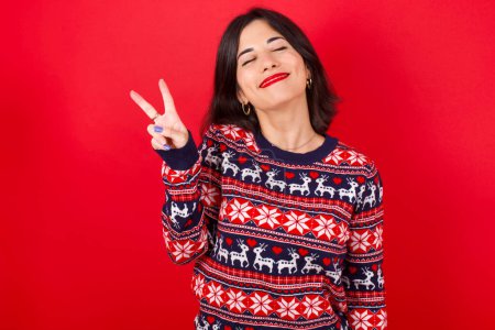 Foto de Morena mujer caucásica vistiendo suéter de Navidad sobre fondo rojo sonriendo con la cara feliz guiñando el ojo a la cámara haciendo signo de victoria. Número dos.. - Imagen libre de derechos
