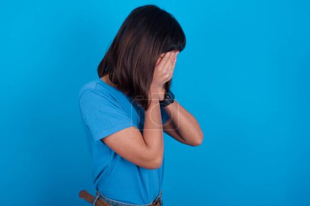 Photo for Sad young asian woman wearing blue t-shirt against blue background covering face with hands and crying. - Royalty Free Image