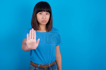 Photo for Young asian woman wearing blue t-shirt against blue background shows stop sign prohibition symbol keeps palm forward to camera with strict expression - Royalty Free Image