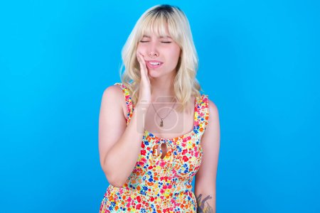 Foto de Concepto de dolor de muelas. Indoor shot of young caucasian girl wearing floral dress isolated over blue background feeling pain, holding his cheek with hand, suffering of bad dentellache, looking at camera with painful expression - Imagen libre de derechos