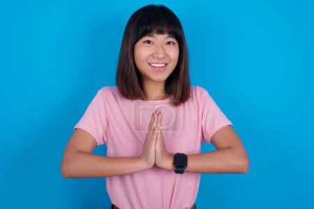 Photo for Young asian woman wearing pink t-shirt against blue background keeps palms together, has pleased expression. Glad attractive male makes request, pleads for mercy. Hopeful young adult. - Royalty Free Image