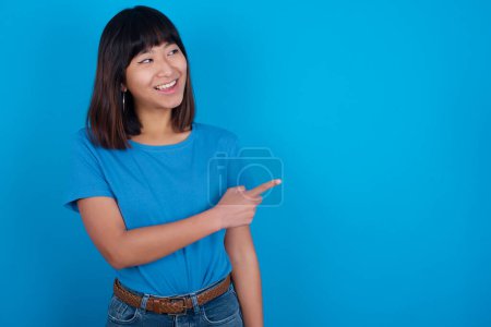Téléchargez les photos : Jeune asiatique femme porter bleu t-shirt sur fond bleu heureux gai démontrant copie espace regarder nouveauté - en image libre de droit