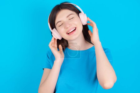 Téléchargez les photos : Jeune fille caucasienne portant bleu t-shirt isolé sur fond bleu studio sourit largement se sent très heureux écoute piste de musique préférée via casque sans fil ferme les yeux. - en image libre de droit