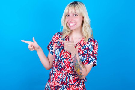 Photo for Optimistic caucasian girl wearing floral dress isolated over blue background points with both hands and  looking at empty space. - Royalty Free Image