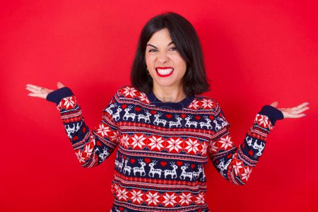 Téléchargez les photos : Clueless femme caucasienne brune portant un pull de Noël sur fond rouge hausse les épaules avec hésitation, fait face à une situation douteuse, écarte les paumes, Décision difficile - en image libre de droit