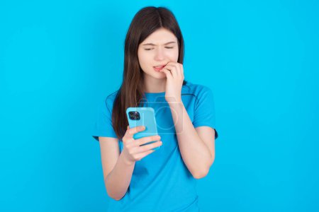 Photo for Portrait of pretty frightened young caucasian girl wearing blue t-shirt isolated over blue studio background chatting biting nails after reading some scary news on her smartphone. - Royalty Free Image