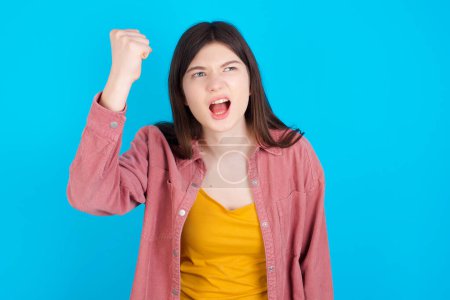 Foto de Chica caucásica joven feroz con camisa rosa aislada sobre fondo azul sosteniendo el puño en frente como si estuviera listo para la pelea o el desafío, gritando y teniendo expresión agresiva en la cara. - Imagen libre de derechos