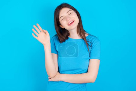 Photo for Overjoyed successful young caucasian girl wearing blue t-shirt isolated over blue studio background raises palm and closes eyes in joy being entertained by friends - Royalty Free Image