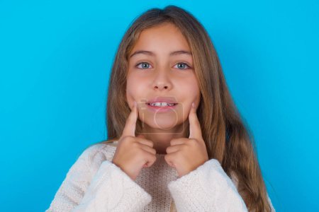 Foto de Feliz chica adolescente bonita con sonrisa dentada, mantiene los dedos índice cerca de la boca, dedos señalando y forzando sonrisa alegre - Imagen libre de derechos