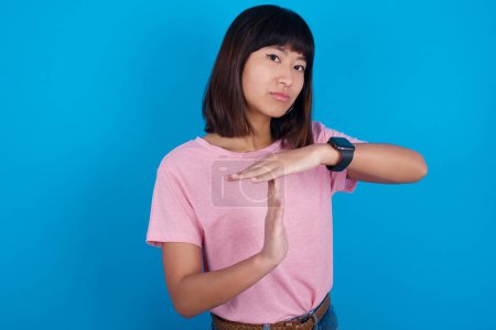 Foto de Joven mujer asiática vistiendo rosa camiseta contra fondo azul se siente cansado y aburrido, haciendo un gesto de tiempo de espera, tiene que parar debido al estrés laboral, concepto de tiempo. - Imagen libre de derechos