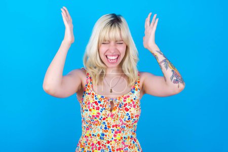 Photo for Caucasian girl wearing floral dress isolated over blue background goes crazy as head goes around feels stressed because of horrible situation - Royalty Free Image
