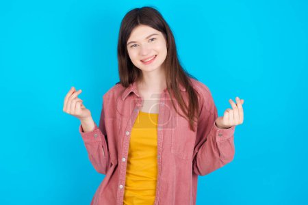 Téléchargez les photos : Jeune fille caucasienne portant chemise rose isolé sur fond bleu faisant geste d'argent. - en image libre de droit