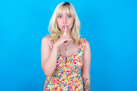 Photo for Surprised caucasian girl wearing floral dress isolated over blue background makes silence gesture, keeps finger over lips and looks mysterious at camera - Royalty Free Image