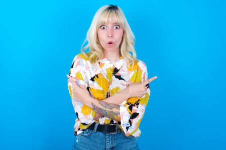 Photo for Confused caucasian girl wearing floral shirt isolated over blue background chooses between two ways, points at both sides with crossed hands, feels doubt. Need your advice. - Royalty Free Image