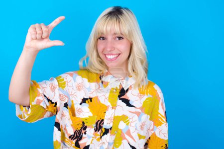 Photo for Caucasian girl wearing floral shirt isolated over blue background smiling and gesturing with hand small size, measure symbol. - Royalty Free Image