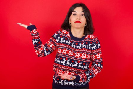 Téléchargez les photos : Brune caucasienne portant un pull de Noël sur fond rouge pointant de côté avec les deux mains montrant quelque chose d'étrange et disant : Je ne sais pas ce que c'est. Concept de publicité. - en image libre de droit