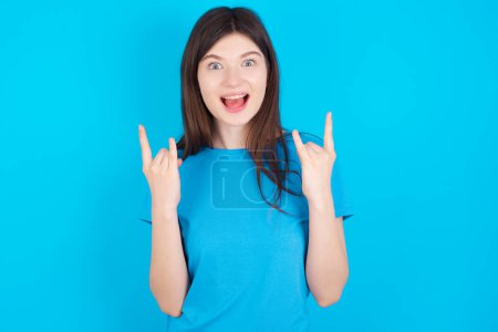 Photo for Young caucasian girl wearing blue t-shirt isolated over blue studio background  makes rock n roll sign looks self confident and cheerful enjoys cool music at party. Body language concept. - Royalty Free Image