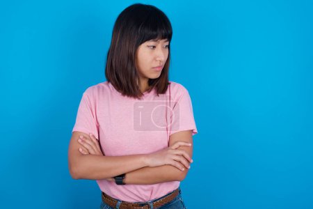Photo for Displeased young asian woman wearing pink t-shirt against blue background with bad attitude, arms crossed looking sideways. Negative human emotion facial expression feelings. - Royalty Free Image