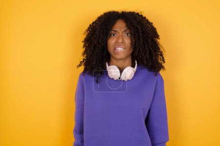 Téléchargez les photos : Portrait de jeune femme afro-américaine stressée avec écouteurs sur fond jaune - en image libre de droit