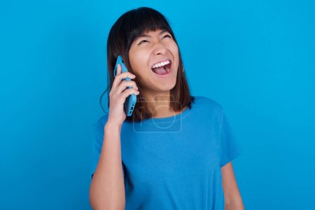 Foto de Overemotive feliz joven asiático mujer usando azul camiseta contra azul fondo risas fuera positivamente oye divertida historia de amigo durante conversación telefónica - Imagen libre de derechos
