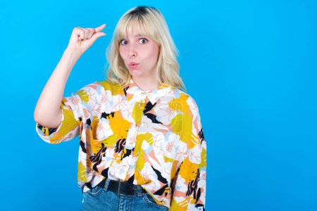 Photo for Shocked Caucasian girl wearing floral shirt isolated over blue background shows something little with hands, demonstrates size, opens mouth from surprise. Measurement concept. - Royalty Free Image