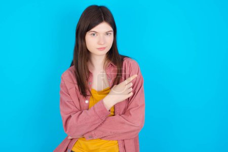 Photo for Young caucasian girl wearing pink shirt isolated over blue background smiling broadly at camera, pointing fingers away, showing something interesting and exciting. - Royalty Free Image