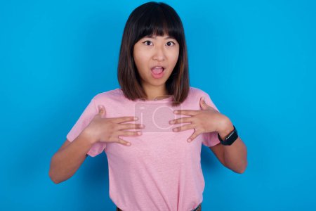 Téléchargez les photos : Jeune femme asiatique portant un t-shirt rose sur fond bleu garde les mains sur la poitrine se sentant choqué et effrayé, la bouche largement ouverte, regarde la caméra en disant : Qui, moi? - en image libre de droit
