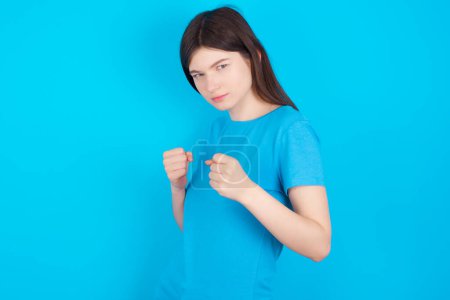 Téléchargez les photos : Portrait de jolie jeune fille caucasienne portant t-shirt bleu isolé sur fond bleu studio tenant la main devant lui en position de boxe va se battre. - en image libre de droit