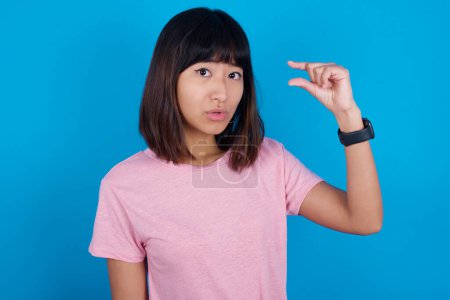 Foto de Joven mujer asiática impactada vistiendo camiseta rosa contra fondo azul muestra algo poco con las manos, demuestra el tamaño, abre la boca de sorpresa. Concepto de medición. - Imagen libre de derechos