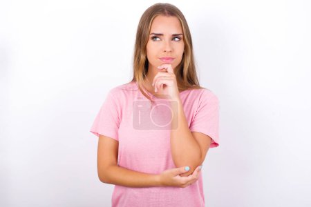 Téléchargez les photos : Pensée jeune fille caucasienne portant T-shirt rose sur fond blanc tient menton et regarde loin pénalement fait grand plan - en image libre de droit